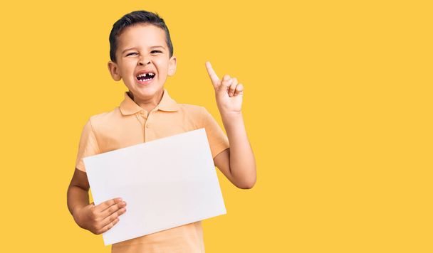 Little cute boy kid holding cardboard banner with blank space surprised with an idea or question pointing finger with happy face, number one  - Foto, Imagen