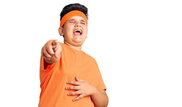 Kleine jongen draagt sportkleding die je uitlacht, wijzend met de vinger naar de camera met hand over lichaam, schaamte uitdrukking  - Foto, afbeelding