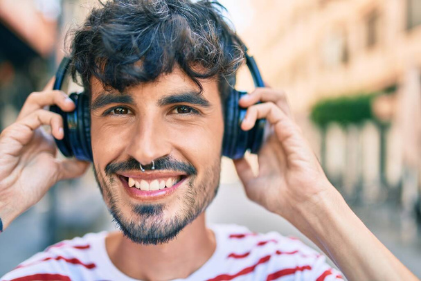 Молодий іспаномовний чоловік посміхається щасливим і використовує навушники, що йдуть вулицею міста
. - Фото, зображення