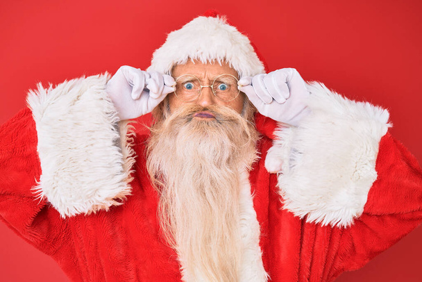 Old senior man with grey hair and long beard wearing santa claus costume holding glasses puffing cheeks with funny face. mouth inflated with air, catching air.  - Photo, Image