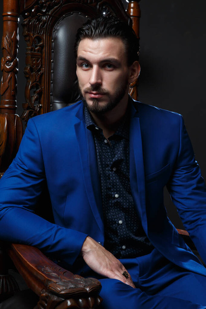 Elegant man in a classic blue suit sitting in a carved wooden chair against a dark background. - Fotoğraf, Görsel
