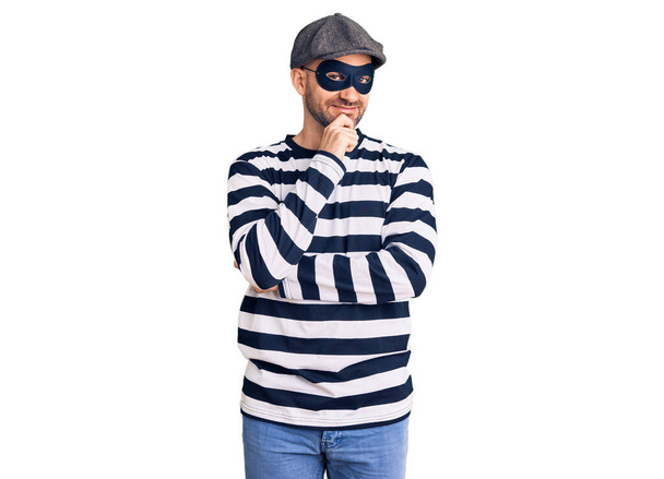 Ein junger gutaussehender Mann mit Einbrechermaske blickt selbstbewusst in die Kamera und lächelt mit verschränkten Armen und erhobener Hand am Kinn. Positives Denken.  - Foto, Bild