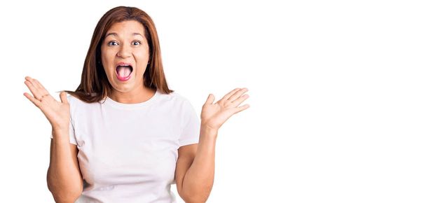 Mujer latina de mediana edad vistiendo una camiseta blanca casual celebrando una locura y sorprendida por el éxito con los brazos levantados y los ojos abiertos gritando emocionados. concepto ganador  - Foto, Imagen