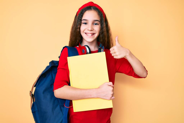 Χαριτωμένο ισπανόφωνο κορίτσι παιδί φορώντας σακίδιο μαθητή και ακουστικά κρατώντας βιβλίο χαμογελώντας χαρούμενος και θετικός, αντίχειρας επάνω κάνει εξαιρετική και την έγκριση σημάδι  - Φωτογραφία, εικόνα