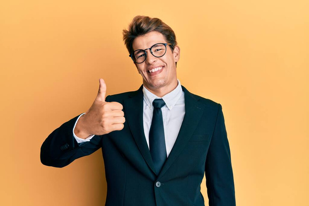 Handsome caucasian man wearing business suit and tie doing happy thumbs up gesture with hand. approving expression looking at the camera showing success.  - 写真・画像