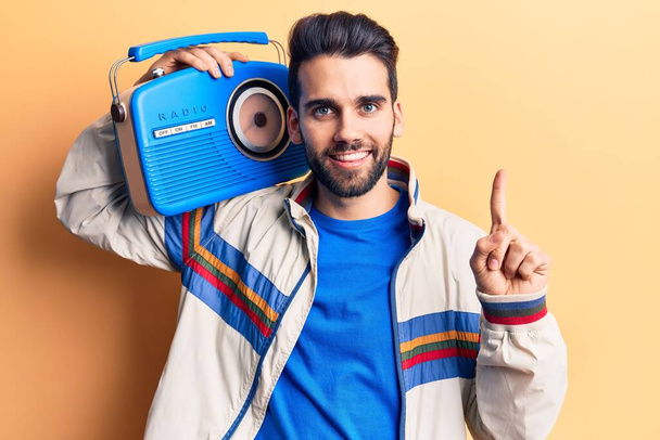 Young handsome man with beard listening to music using vintage boombox smiling with an idea or question pointing finger with happy face, number one  - Photo, Image