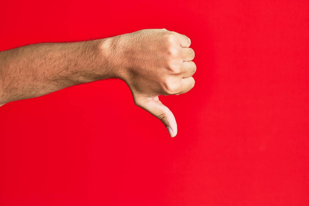 Braço de branco branco jovem sobre fundo vermelho isolado fazendo polegares para baixo gesto de rejeição, desaprovação antipatia e sinal negativo  - Foto, Imagem