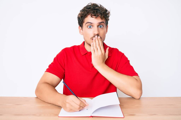 Młody biały mężczyzna z kręconymi włosami, piszący notatnik, siedzący na stole i zakrywający usta ręką, zszokowany i przerażony pomyłką. zaskoczona ekspresja  - Zdjęcie, obraz
