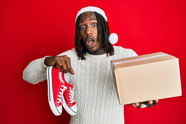 Afro-Amerikaanse man met vlechten dragen kerstmuts met schoenen als een geschenk in shock gezicht, op zoek sceptisch en sarcastisch, verrast met open mond  - Foto, afbeelding