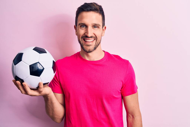 Fiatal jóképű játékos férfi focizni tartó labdarúgó labda felett elszigetelt rózsaszín háttér néz pozitív és boldog álló és mosolygós magabiztos mosollyal mutatja fogak - Fotó, kép