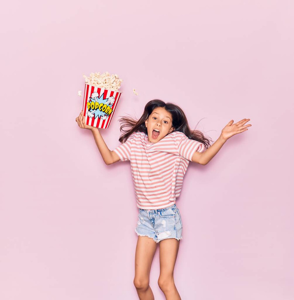 Adorable hispanic child girl surprised with open mouth holding bucket of popcorn jumping over isolated pink background - Фото, зображення