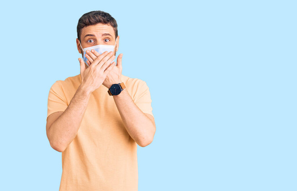 Schöner junger Mann mit Bär mit medizinischer Maske für Coronavirus schockiert Mund mit Händen für Fehler bedecken. Geheimes Konzept.  - Foto, Bild