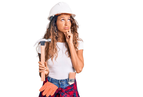 Young hispanic woman with tattoo wearing hardhat and builder clothes holding hammer serious face thinking about question with hand on chin, thoughtful about confusing idea  - Fotó, kép