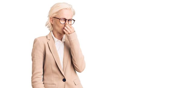 Young blonde woman wearing business clothes and glasses smelling something stinky and disgusting, intolerable smell, holding breath with fingers on nose. bad smell  - Fotoğraf, Görsel