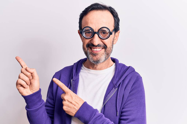 Middle age handsome freaky man wearing dumb glasses over isolated white background smiling and looking at the camera pointing with two hands and fingers to the side. - Foto, Imagen