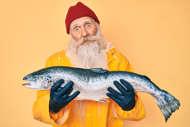 Homem idoso com cabelos grisalhos e barba comprida vestindo capa de chuva segurando salmão fresco no rosto de choque, parecendo cético e sarcástico, surpreso com a boca aberta  - Foto, Imagem