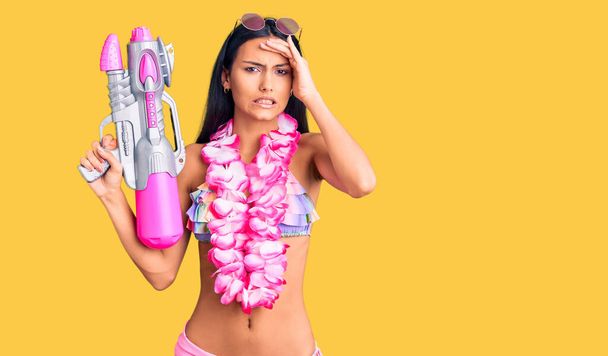 Fiatal gyönyörű latin lány visel bikini és hawaii lei tartja vízipisztoly stresszes és frusztrált kézzel a fejét, meglepett és dühös arc  - Fotó, kép