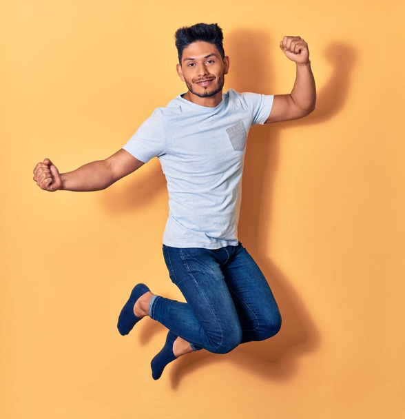 Young handsome latin man wearing casual clothes smiling happy. Jumping with smile on face doing winner sign with fists up over isolated yellow background - Valokuva, kuva