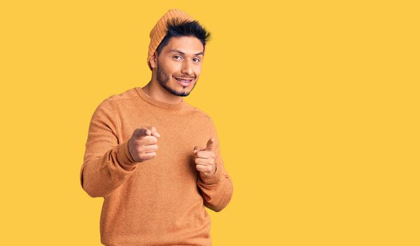 冬のセーターとウールの帽子を身に着けているハンサムなラテン系アメリカ人の若者は、幸せで面白い顔をしてカメラに指を指しています。良いエネルギーと振動は.  - 写真・画像