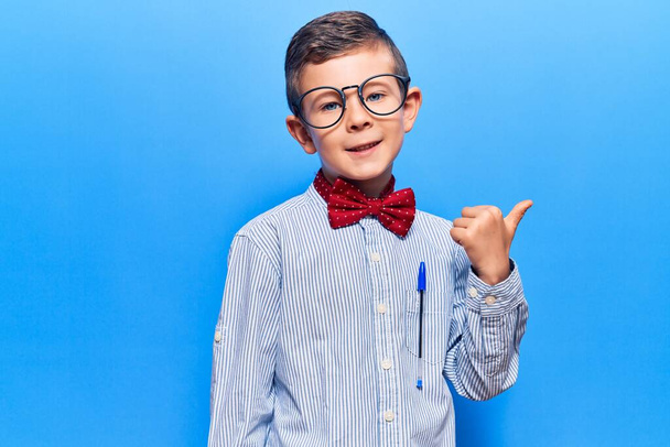 Lindo niño rubio con corbata de lazo nerd y gafas sonriendo con la cara feliz mirando y señalando a un lado con el pulgar hacia arriba.  - Foto, Imagen