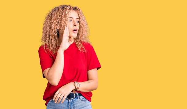 Young blonde woman with curly hair wearing casual red tshirt hand on mouth telling secret rumor, whispering malicious talk conversation  - Photo, Image
