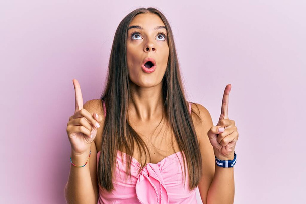 Young hispanic woman wearing casual style with sleeveless shirt amazed and surprised looking up and pointing with fingers and raised arms.  - Foto, Imagem
