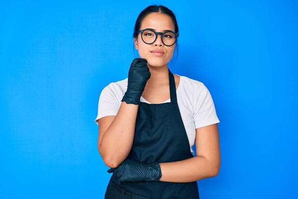 Young beautiful asian girl wearing professional apron serious face thinking about question with hand on chin, thoughtful about confusing idea  - Photo, Image