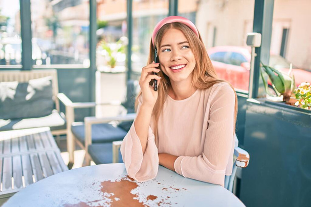 Junges kaukasisches Mädchen lächelt glücklich und spricht auf der Café-Terrasse auf dem Smartphone. - Foto, Bild