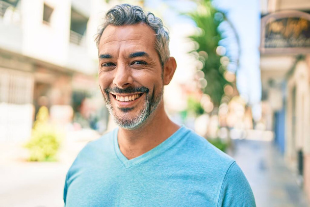 Hombre de pelo gris de mediana edad sonriendo feliz caminando en la calle de la ciudad. - Foto, imagen