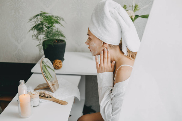 Young woman in white towel chilling in bedroom and making clay facial mask near mirror. Girl doing beauty treatment and relaxing at home. Morning skin care beauty routine, self care. - Photo, Image