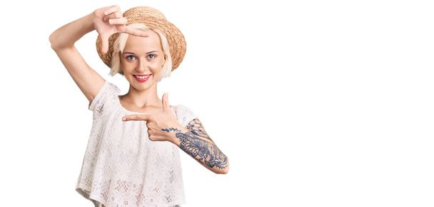 Mujer rubia joven con tatuaje con sombrero de verano sonriente haciendo marco con las manos y los dedos con la cara feliz. concepto de creatividad y fotografía.  - Foto, imagen