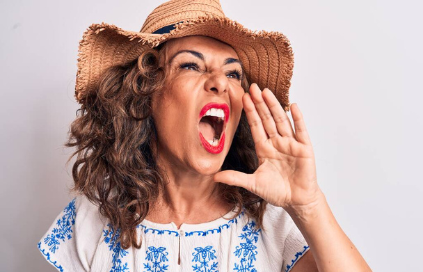 Středního věku krásná brunetka žena na dovolené na sobě letní klobouk přes bílé pozadí křičí a křičí hlasitě na stranu s rukou na ústech. Koncept komunikace. - Fotografie, Obrázek