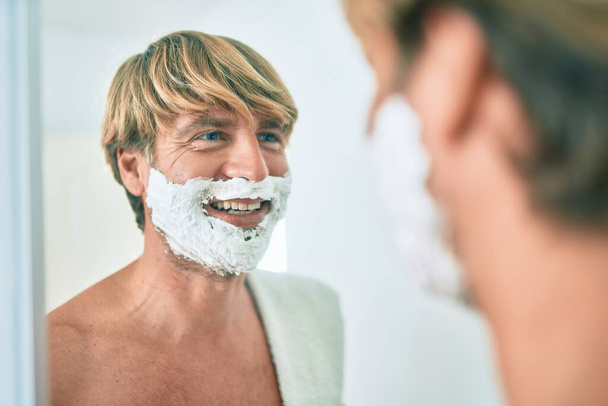 Hombre rubio guapo mirando el espejo en el baño ahorro usando espuma y afeitadora - Foto, imagen