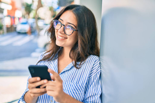 Junge hispanische Geschäftsfrau lächelt glücklich mit Smartphone in die Stadt. - Foto, Bild