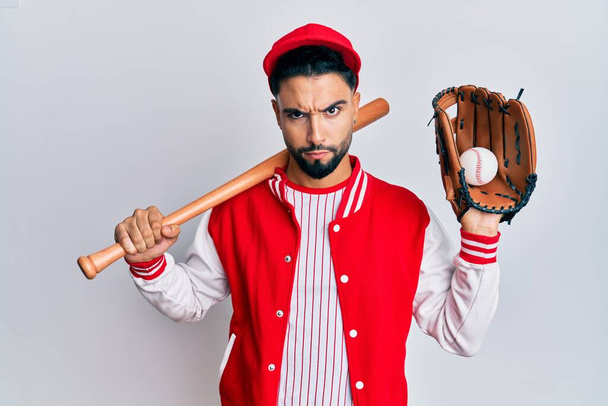 Молодой человек с бородой играет в бейсбол держа летучую мышь и мяч скептически и нервно, хмурясь расстроен из-за проблемы. отрицательный человек.  - Фото, изображение