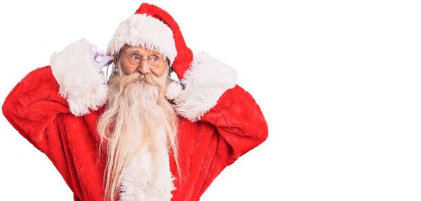 Starý starší muž s šedivými vlasy a dlouhými vousy v tradičním kostýmu Santa Clause, s úsměvem tahající za uši prsty, legrační gesto. problém konkurzu  - Fotografie, Obrázek