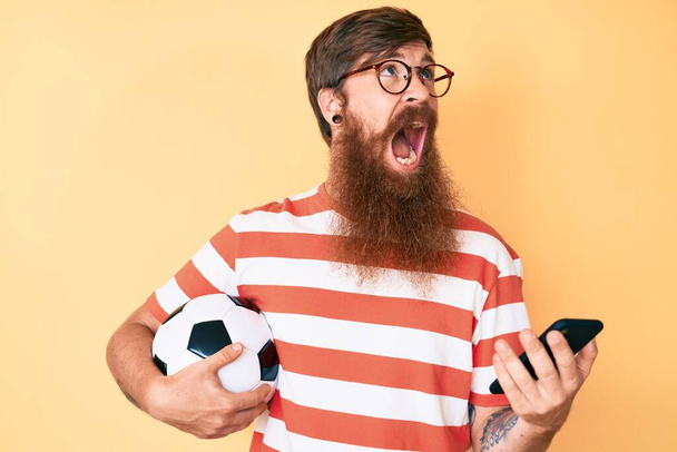 Guapo joven cabeza roja hombre con barba larga sosteniendo pelota de fútbol mirando teléfono inteligente enojado y loco gritando frustrado y furioso, gritando con ira mirando hacia arriba.  - Foto, imagen