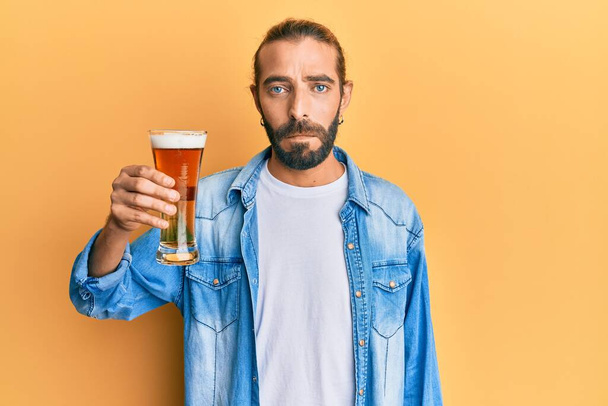 Homem atraente com cabelos longos e barba bebendo uma caneca de cerveja pensando atitude e expressão sóbria olhando auto confiante  - Foto, Imagem