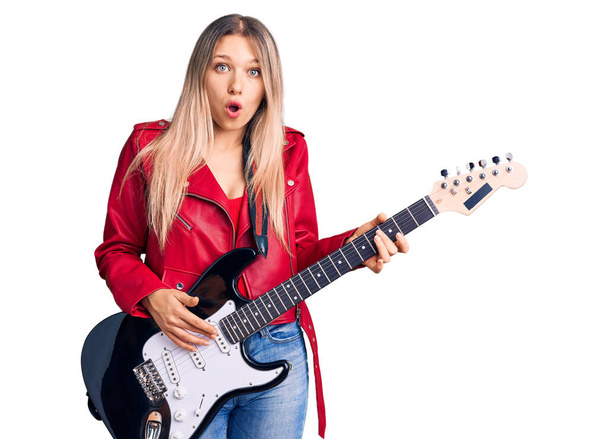Молода красива блондинка грає на електричній гітарі налякана і вражена відкритим ротом для несподіванки, обличчя віри
  - Фото, зображення