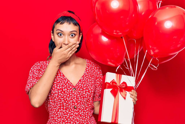 Mladá žena držící balónky s dárkem zakrývajícím ústa rukou, šokovaná a vystrašená z omylu. překvapený výraz  - Fotografie, Obrázek