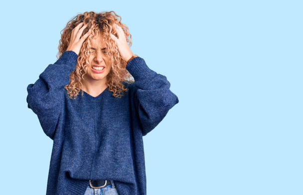 Jonge blonde vrouw met krullend haar draagt casual winter trui die lijdt aan hoofdpijn wanhopig en gestrest omdat pijn en migraine. handen op het hoofd.  - Foto, afbeelding