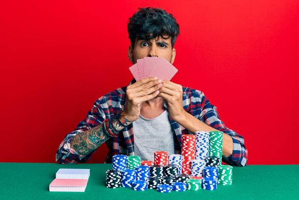 Mladý hispánec hrající poker zakrývající tvář s kartami v šoku, vypadá skepticky a sarkasticky, překvapen otevřenými ústy  - Fotografie, Obrázek