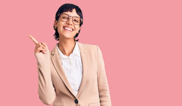 Junge brünette Frau mit kurzen Haaren trägt Businessjacke und Brille und zeigt mit Finger Nummer eins nach oben, während sie selbstbewusst und glücklich lächelt.  - Foto, Bild