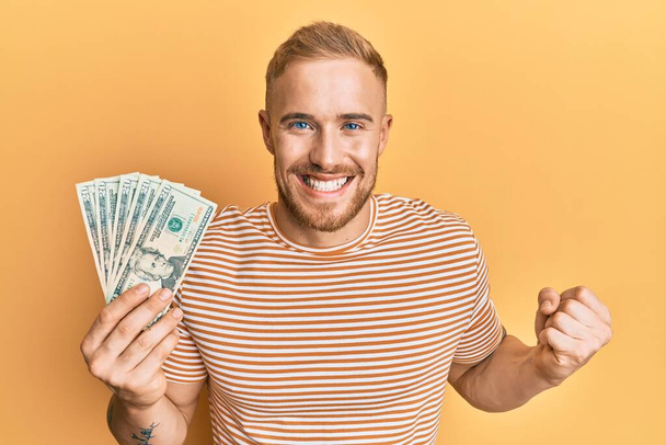 Young caucasian man holding dollars screaming proud, celebrating victory and success very excited with raised arm  - Photo, Image