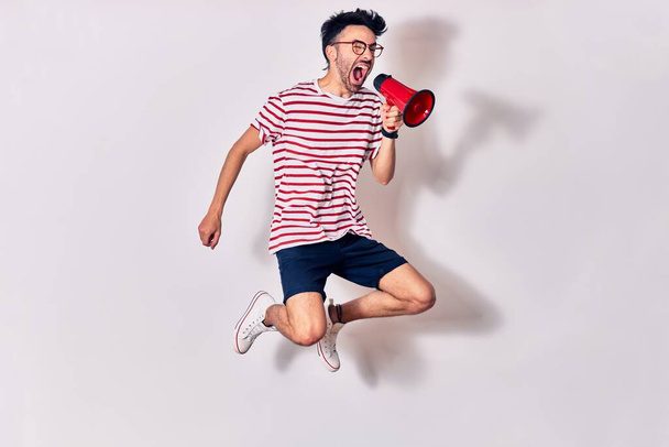 Joven hombre hispano guapo con gafas gritando usando megáfono. Saltar sobre fondo blanco aislado - Foto, Imagen