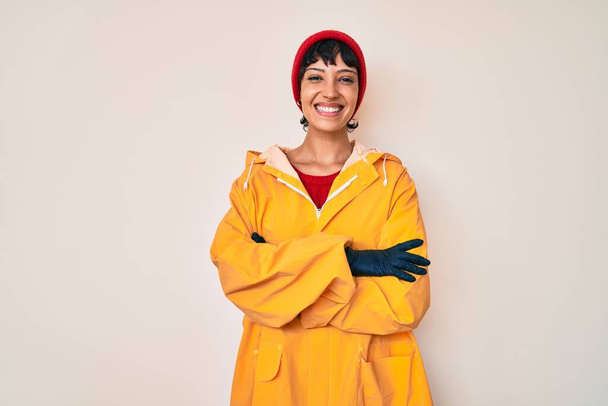 Beautiful brunettte woman wearing yellow raincoat happy face smiling with crossed arms looking at the camera. positive person.  - Фото, изображение