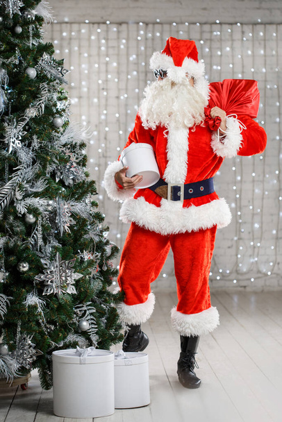 A man in a Santa Claus costume with a bag of gifts near a Christmas tree in a bright room. - Fotó, kép