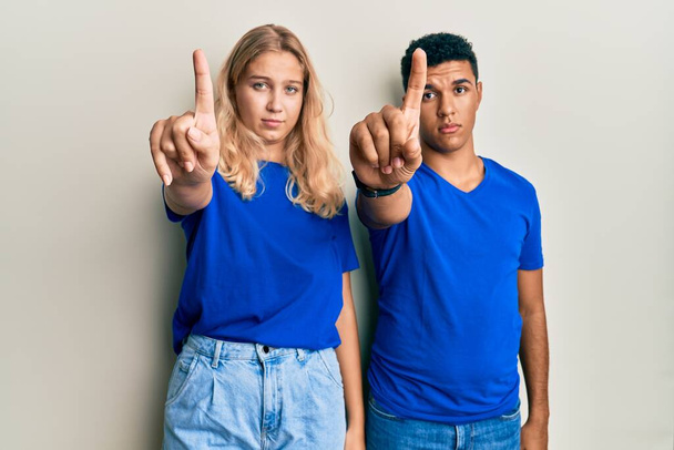 Νεαρό διαφυλετικό ζευγάρι που φοράει καθημερινά ρούχα δείχνοντας με το δάχτυλο προς τα πάνω και θυμωμένη έκφραση, χωρίς να δείχνει καμία χειρονομία  - Φωτογραφία, εικόνα