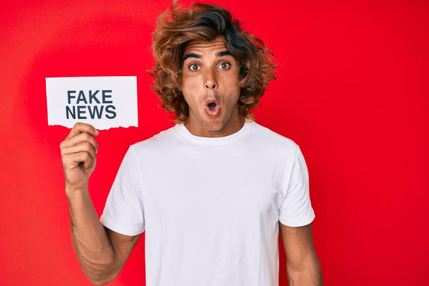 Joven hombre hispano sosteniendo pancarta de noticias falsas asustado y sorprendido con la boca abierta para sorpresa, cara de incredulidad  - Foto, Imagen