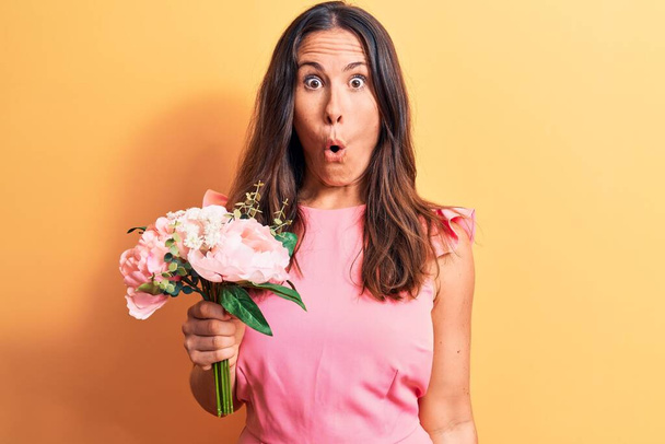 Young beautiful brunette woman holding bouquet of pink flowers over yellow background scared and amazed with open mouth for surprise, disbelief face - Foto, Bild